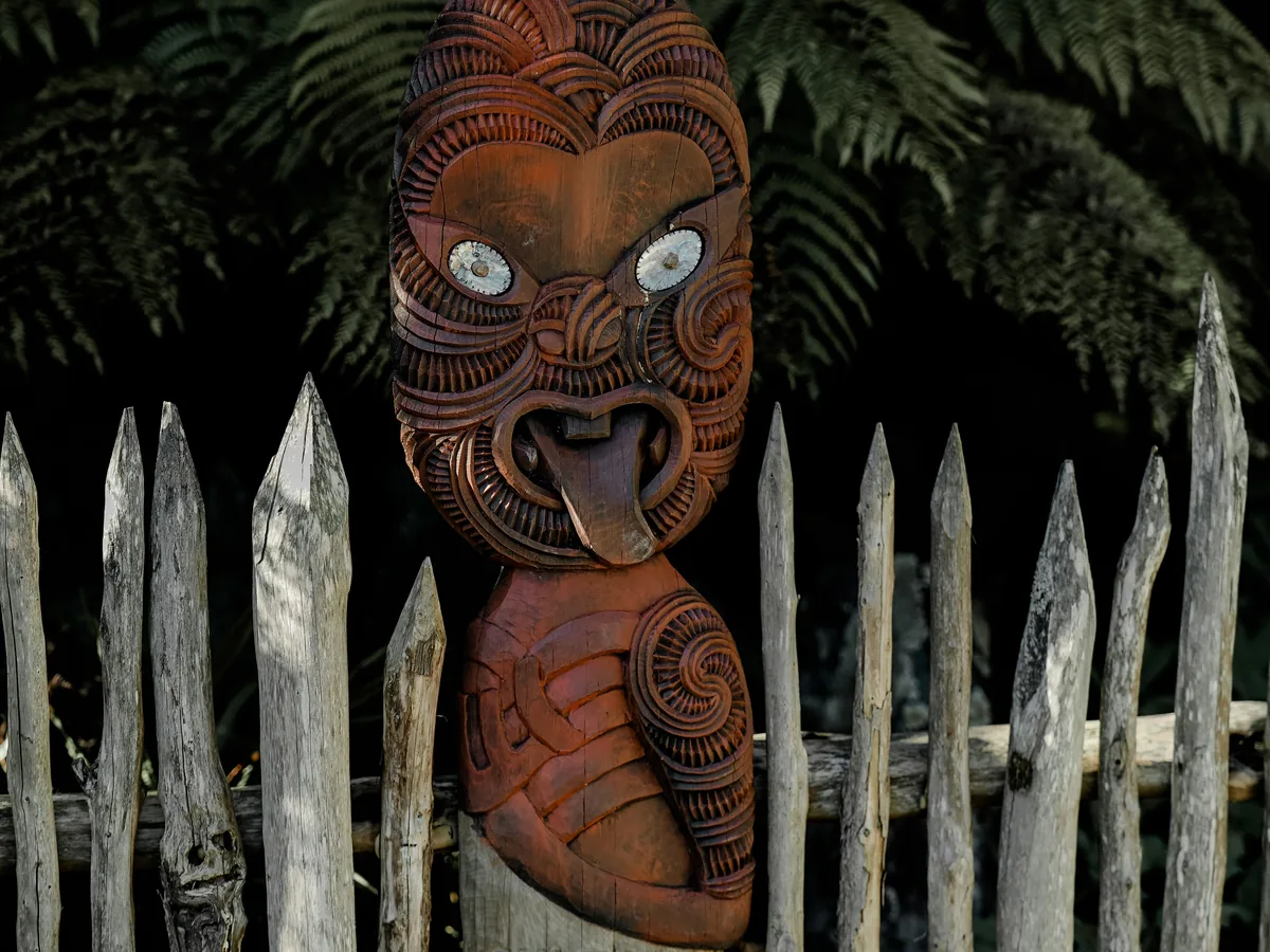 A carved wooden Māori guardian figure set into a palisade fence among tree ferns — representative carving, not Te Puia's own