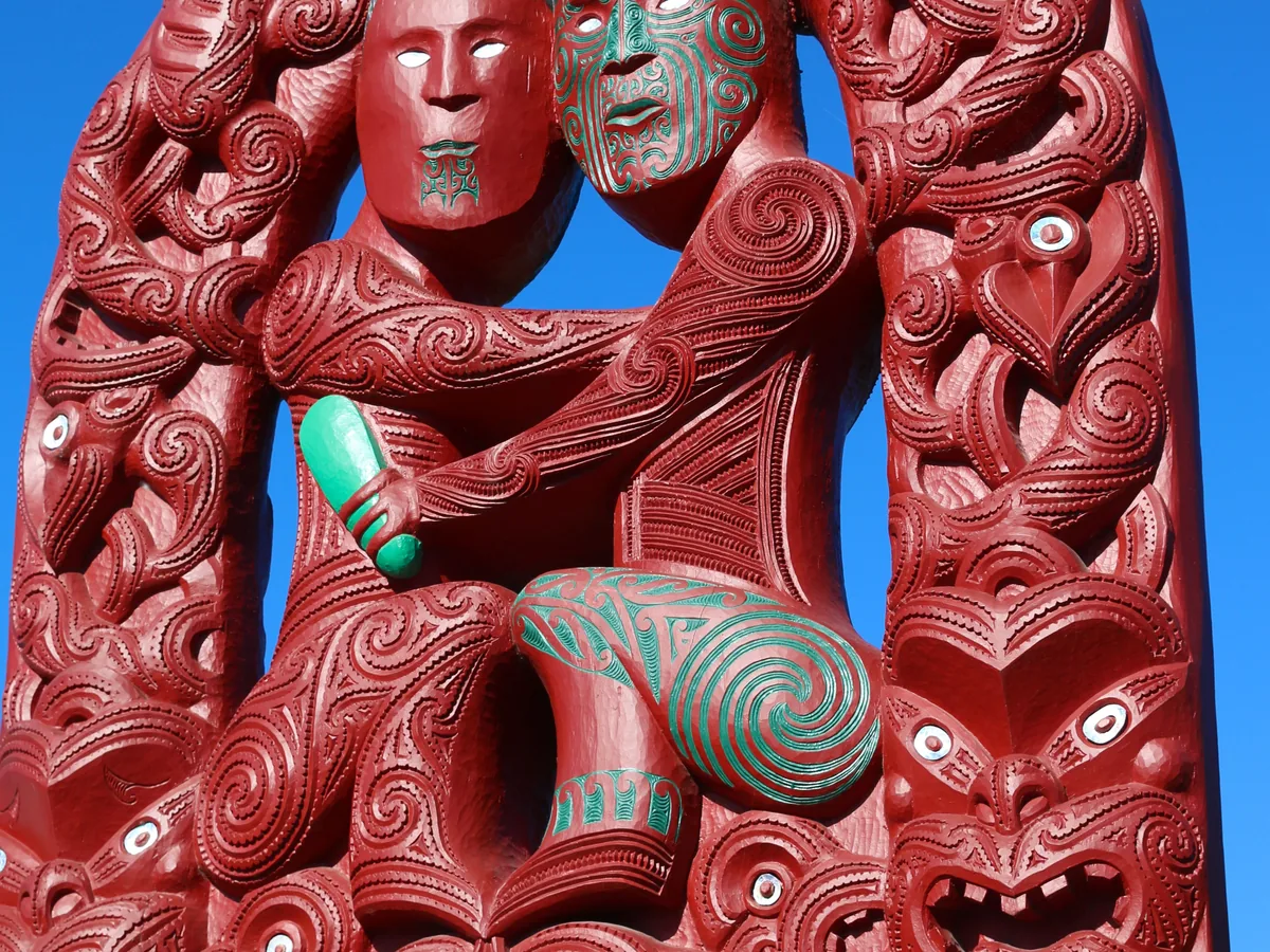 A carved red and pounamu-green Māori gateway figure against a blue sky — representative carving, not photographed at Te Puia