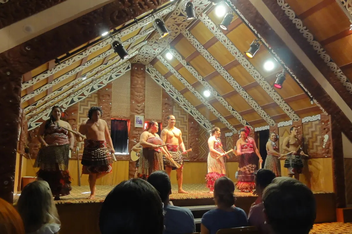 Performers in woven skirts and cloaks on a small stage inside a carved wooden meeting house, lit by spotlights, with an audience in the foreground