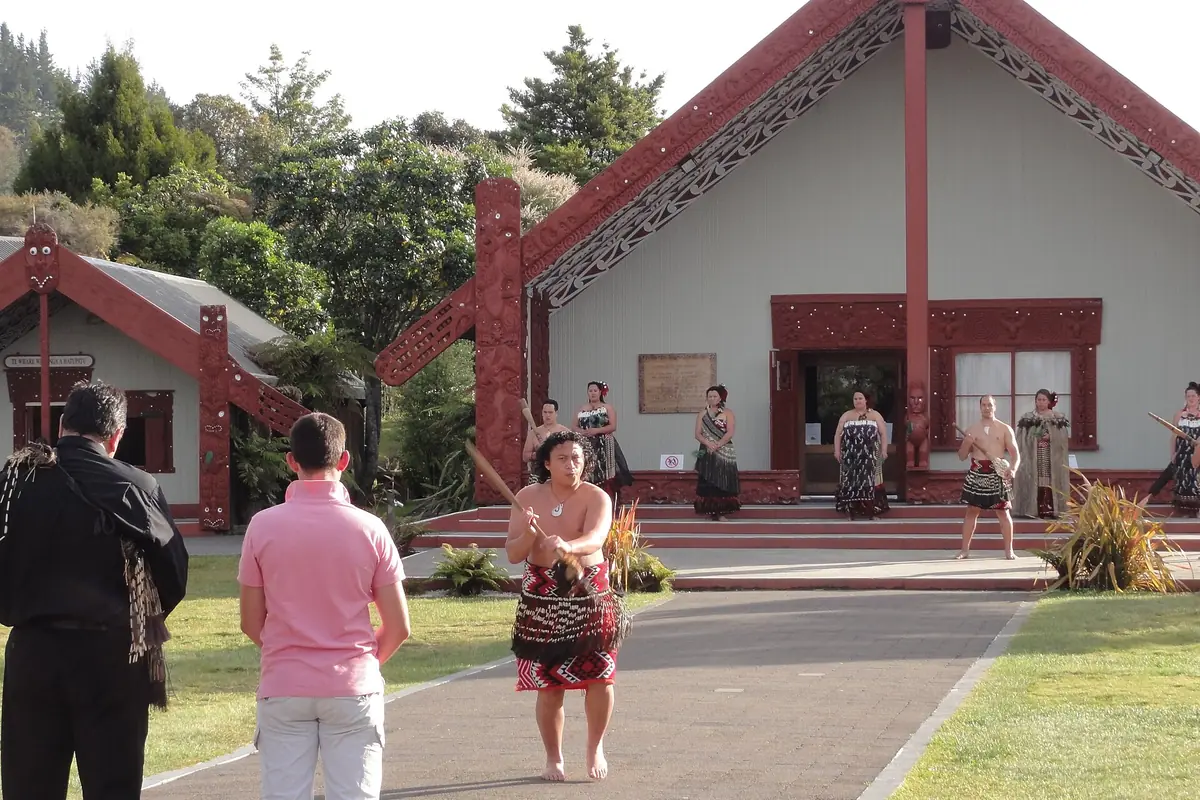 A performer with a taiaha stands on a path in front of a carved red and white meeting house at Te Puia, with other performers on the porch behind